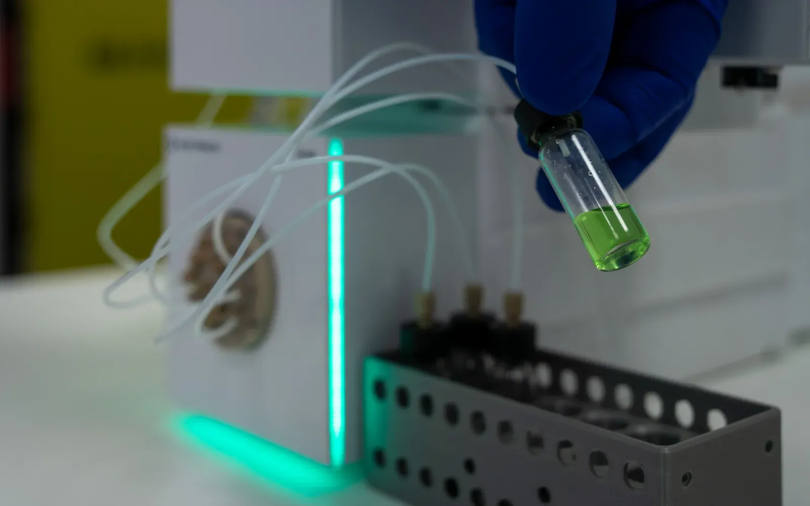 A gloved hand placing a vial in the QB AutoSampler vial rack next to the Selector Valve